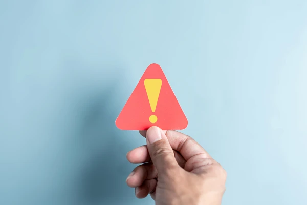 Hand holding up paper cutout of red triangle with a yellow exclamation point against a light blue backdrop