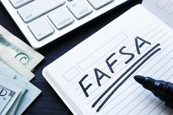 A notebook with "FAFSA" written on it sits next to a stack of cash and a computer keyboard.