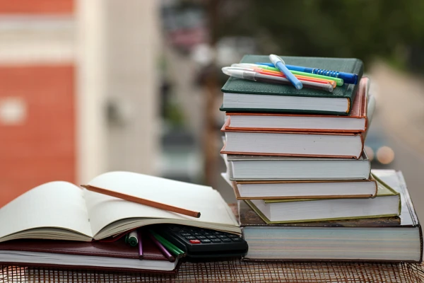 A stack of books topped with pens next to an open notebook, pens, and a calculator on an outdoor table.