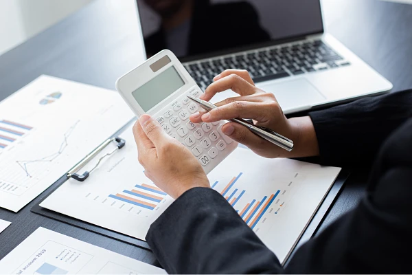 A person is using a calculator at a desk with financial charts and a laptop.