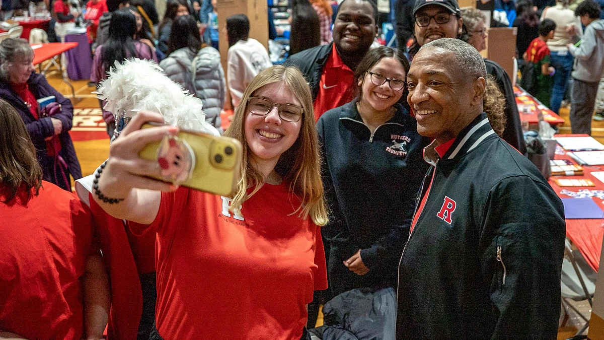 A group of people in red attire, including one in a Rutgers jacket, take a selfie together at an indoor event.