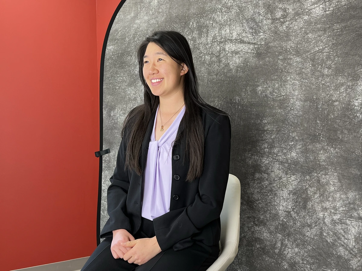 A woman in a black blazer is sitting and smiling against a textured gray backdrop in a photo studio setting.